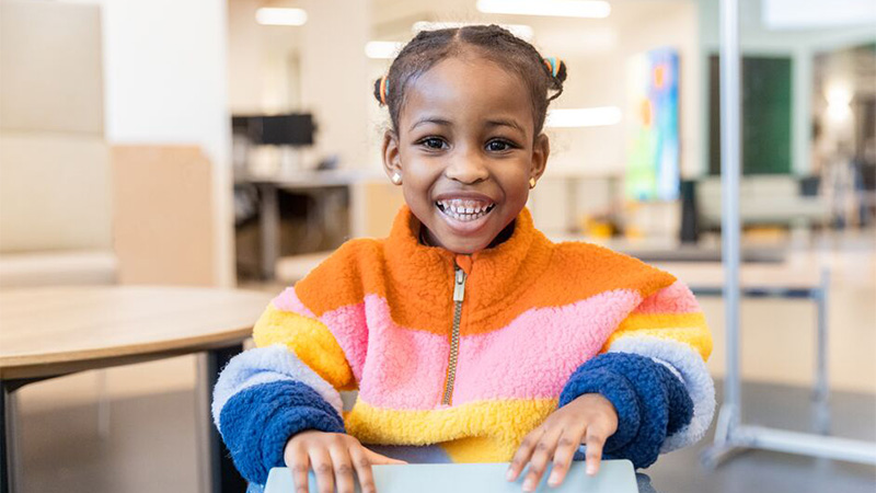 A younger school-aged girl with braided hair wearing a rainbow quarter zip fleece smiles directly at the camera.