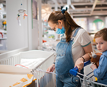 Mother looks at a crib while her young son sits in the shopping cart beside her